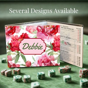 May include: A floral-patterned card holder with the name "Debbie" on a pink label. The design features red and pink peonies. The text "Several Designs Available" is at the top. Dice are scattered on a green surface.