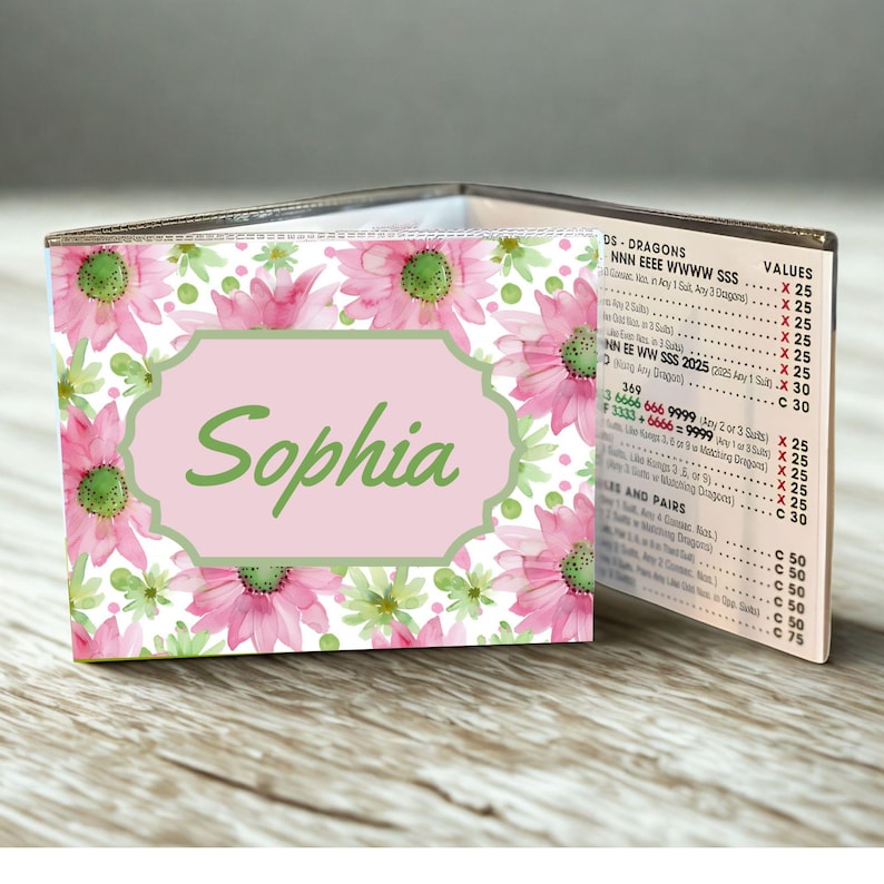May include: A Mah Jongg card holder featuring a watercolor floral pattern of pink and green flowers on a white background. A central light pink label with a green border displays the name Sophia in green script. The side shows a printed score chart.