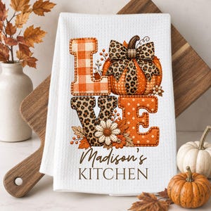 Op de afbeelding: Een witte wafelstructuur keukendoek met het woord LOVE, waarbij een pompoen de O vervangt. De L is oranje geruit, de O is een pompoen met een luipaardprint strik, de V is luipaardprint en de E is oranje met witte stippen. Tekst: Madison's KITCHEN.