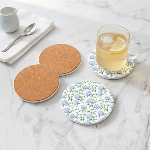 May include: Three round coasters, two with a natural brown cork texture and one featuring a white surface with a repeating pattern of blue hydrangea flowers and green leaves. A glass mug with tea and a lemon slice sits on the floral coaster.