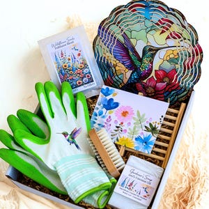 May include: A gardening gift set in a white box. Includes green and white gardening gloves with a hummingbird design, a wooden brush, a decorative hummingbird art piece, wildflower seeds, a floral card, and a bar of Gardener's Soap.
