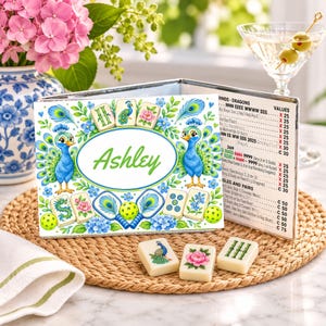 Op de afbeelding: Een Mahjong-kaarthouder met de gepersonaliseerde naam Ashley in groen schrift, omringd door illustraties van blauwe pauwen, bloemen en speelstenen. Een gedrukte scorekaart is aan de zijkant bevestigd, met drie losse stenen ervoor.