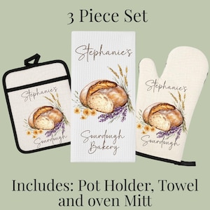 Op de afbeelding: Een driedelige keukenset bestaande uit een vierkante pannenlap, een rechthoekige theedoek en een ovenwant. Elk item is gebroken wit met een print van een brood, tarwe en bloemen, voorzien van de tekst Stephanie's Sourdough Bakery.