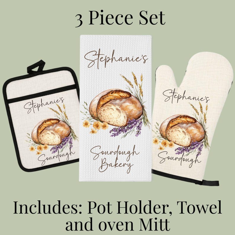Op de afbeelding: Een driedelige keukenset bestaande uit een vierkante pannenlap, een rechthoekige theedoek en een ovenwant. Elk item is gebroken wit met een print van een brood, tarwe en bloemen, voorzien van de tekst Stephanie's Sourdough Bakery.