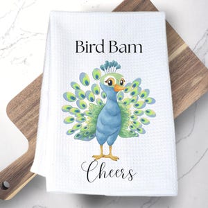 Op de afbeelding: Een witte wafelstructuur keukendoek met een kleurrijke illustratie van een pauw met blauwe, groene en gele veren. De tekst "Bird Bam" is in het zwart boven de pauw gedrukt en het woord "Cheers" is in het zwart eronder gedrukt.