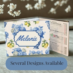 May include: A folded card featuring a pattern of blue hydrangeas and yellow lemons on a cream background. The front displays a central label with the name Melanie in blue script. The interior panel shows a printed list of game values and rules.