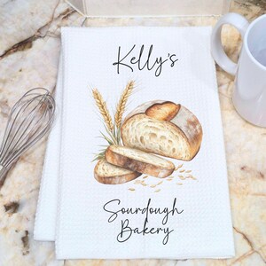 May include: A white waffle-weave kitchen towel featuring a watercolor illustration of a loaf of sourdough bread, a slice, and wheat stalks. The text Kelly's Sourdough Bakery is printed in black script. A metal whisk and a white mug are nearby.