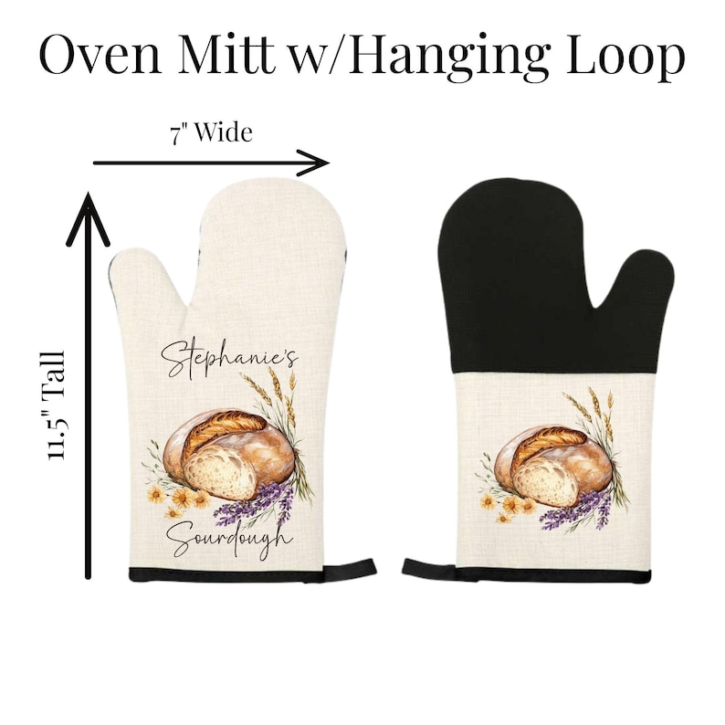 Op de afbeelding: Twee ovenwanten met ophanglussen, 17,78 cm breed en 29,21 cm hoog. De ene is beige met een illustratie van zuurdesembrood, tarwe, bloemen en de tekst 'Stephanie's Sourdough'. De andere heeft een zwarte bovenkant en een beige onderkant.