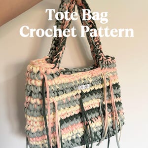 May include: A handmade tote bag with a crochet pattern. The bag features a mix of pink, gray, and cream-colored yarn, with dark green accents on the handles and fringe. The text "Tote Bag Crochet Pattern" is displayed on the image.