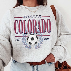 May include: Gray sweatshirt with a graphic design that says "Soccer Colorado Est 1995" with a soccer ball in the center. The sweatshirt has a crew neck and long sleeves.