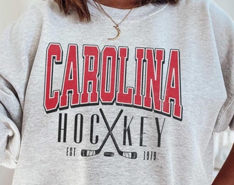 Puede incluir: Sudadera gris con un gráfico rojo de "CAROLINA HOCKEY" con palos de hockey cruzados y el texto "EST 1979".