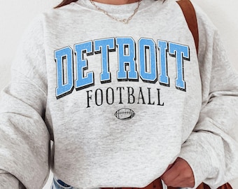 May include: An ash gray crewneck sweatshirt featuring the text DETROIT FOOTBALL in bold light blue letters with black outlines, accompanied by a small black football graphic. The sweatshirt is paired with light blue denim jeans and a brown belt.