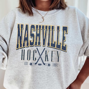 May include: A grey sweatshirt with the text "Nashville Hockey Est 1997" in navy blue and yellow lettering. The sweatshirt features a graphic of two crossed hockey sticks.