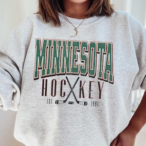 Peut inclure: Sweat-shirt gris avec un motif vert et blanc qui dit "Minnesota Hockey Est 1997".