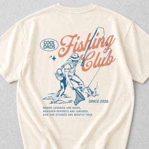 May include: Ivory t-shirt featuring a fishing-themed graphic with the words "Fishing Club" and "Cool Dads." The design includes the text "Since 2026" and a quote. A casual wear item.