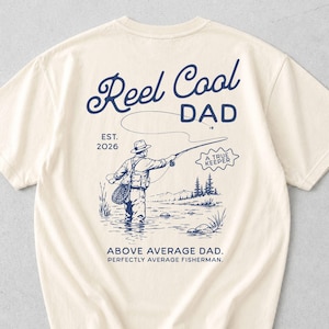 May include: Cream-colored t-shirt with a navy blue graphic on the back. The graphic features the words "Reel Cool Dad" above an illustration of a fisherman. Below the illustration are the words "Above Average Dad. Perfectly Average Fisherman."