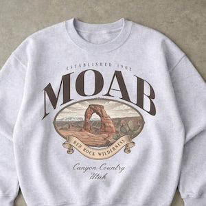 May include: Light gray sweatshirt with a Moab, Utah graphic. The graphic features an illustration of a red rock archway, with the words "MOAB" and "Red Rock Wilderness" in brown lettering. The sweatshirt also has the text "Established 1902" and "Canyon Country Utah".