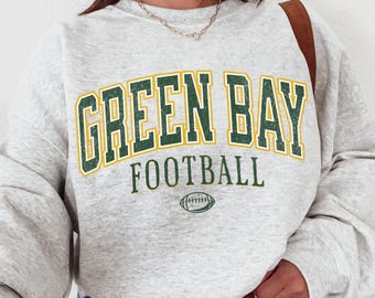 May include: A light gray heathered crewneck sweatshirt featuring the text GREEN BAY in distressed green letters with a yellow outline, the word FOOTBALL in green below it, and a small green football graphic centered at the bottom of the design.
