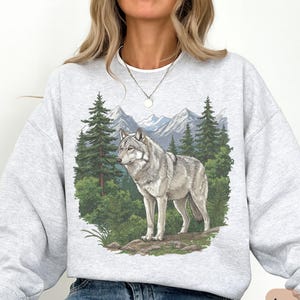 Op de afbeelding: Lichtgrijze sweatshirt met een gedetailleerde illustratie van een grijze wolf in een boslandschap. Het ontwerp omvat groenblijvende bomen, bergen en een natuurlijk landschap. Het woord "ASH" staat rechtsonder gedrukt.
