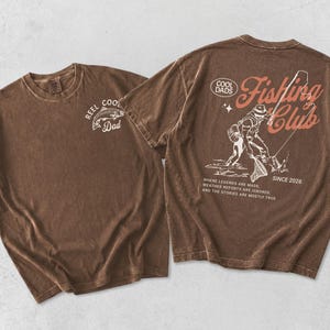 May include: Two brown t-shirts. One has "REEL COOL Dad" and a fish graphic. The other has a fishing scene with text "Fishing Club Since 2026" and "Where legends are made, weather reports are ignored, and the stories are mostly true."