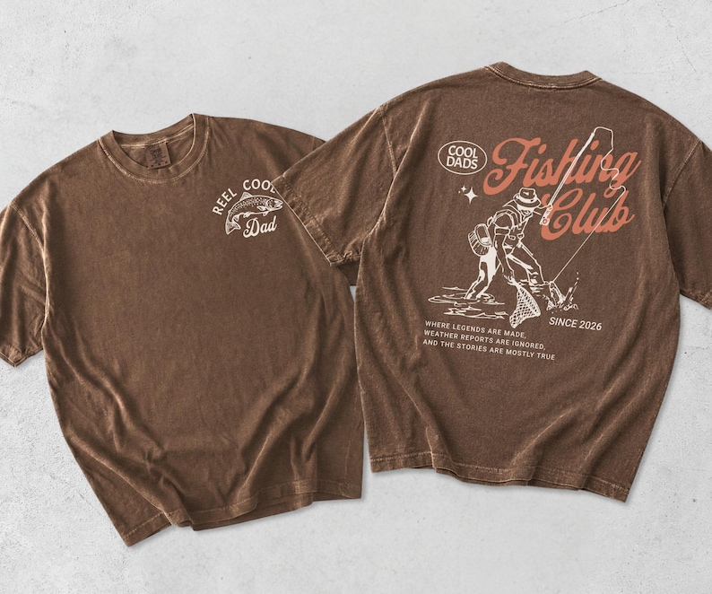 May include: Two brown t-shirts. One has "REEL COOL Dad" and a fish graphic. The other has a fishing scene with text "Fishing Club Since 2026" and "Where legends are made, weather reports are ignored, and the stories are mostly true."