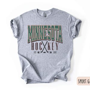 Peut inclure: Un t-shirt gris avec le texte "Minnesota Hockey Est 1967" en lettres vertes, brunes et blanches. Le texte est entour&eacute; de deux b&acirc;tons de hockey crois&eacute;s.