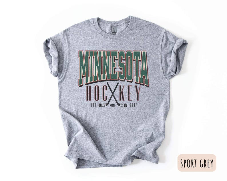 Peut inclure: Un t-shirt gris avec le texte "Minnesota Hockey Est 1967" en lettres vertes, brunes et blanches. Le texte est entour&eacute; de deux b&acirc;tons de hockey crois&eacute;s.