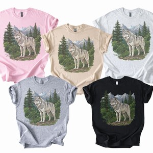 Op de afbeelding: Vijf T-shirts in verschillende kleuren: roze, beige, lichtgrijs, grijs en zwart. Elk shirt heeft een afbeelding van een wolf in een bos met bergen op de achtergrond. De wolf is grijs en wit.