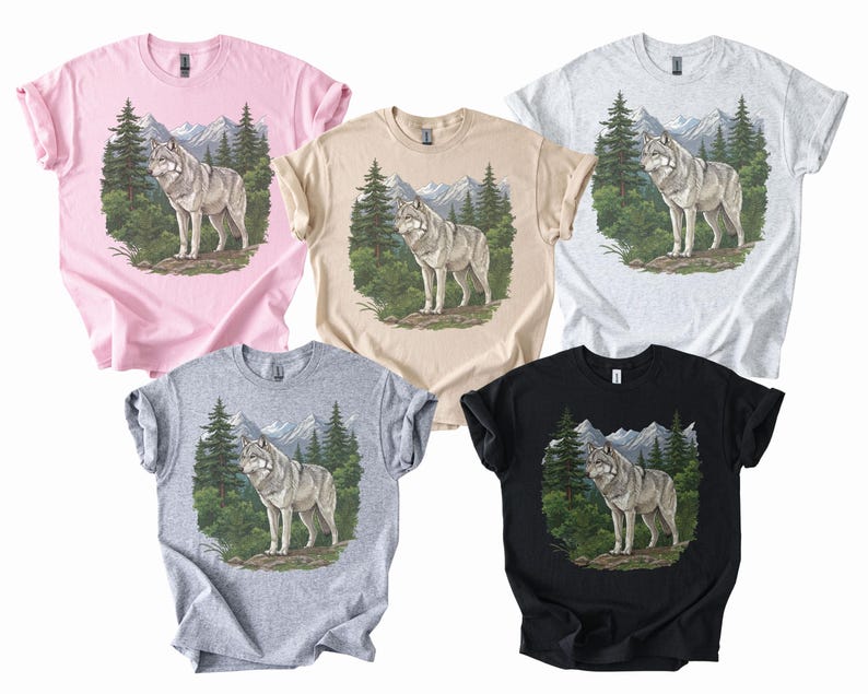 Op de afbeelding: Vijf T-shirts in verschillende kleuren: roze, beige, lichtgrijs, grijs en zwart. Elk shirt heeft een afbeelding van een wolf in een bos met bergen op de achtergrond. De wolf is grijs en wit.