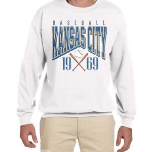 Peut inclure: Sweat-shirt blanc avec un motif bleu et or qui dit "Baseball Kansas City 19 69" avec un ballon de baseball et des chauves-souris crois&eacute;es.