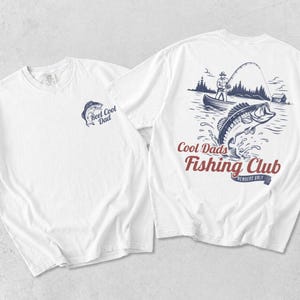 May include: White t-shirts with fishing-themed graphics. One shirt features a blue fish and the text "Reel Cool Dad." The other shirt displays a fisherman in a boat, a jumping fish, and the words "Cool Dads Fishing Club, Members Only."