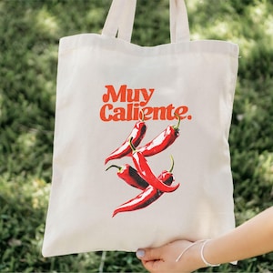 May include: Off-white tote bag featuring the phrase "Muy Caliente" in red script above a graphic of three red chili peppers. The bag has two handles and is held up by a person.
