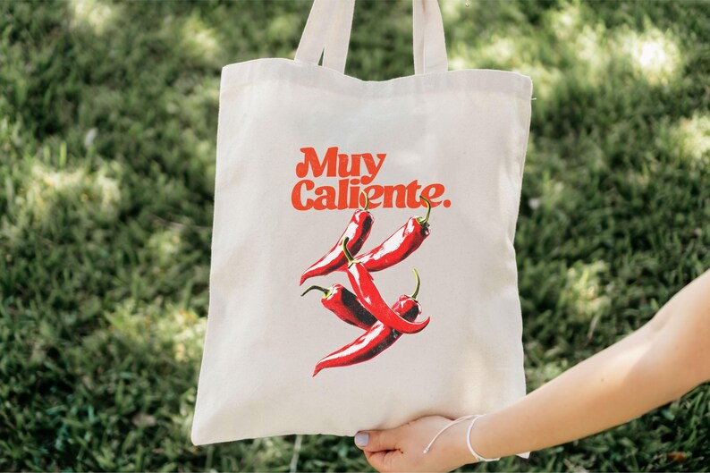 May include: Off-white tote bag featuring the phrase "Muy Caliente" in red script above a graphic of three red chili peppers. The bag has two handles and is held up by a person.