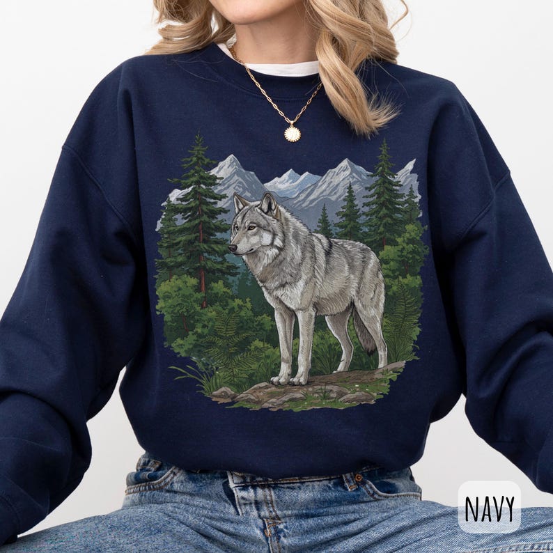 Op de afbeelding: Marineblauwe sweatshirt met een gedetailleerde illustratie van een grijze wolf in een boslandschap met bergen. Het woord "NAVY" is rechtsonder in het wit gedrukt.