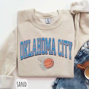 May include: A cream coloured sweatshirt with the text "OKLAHOMA CITY" in blue letters with a basketball and net graphic. The sweatshirt is on a denim background.