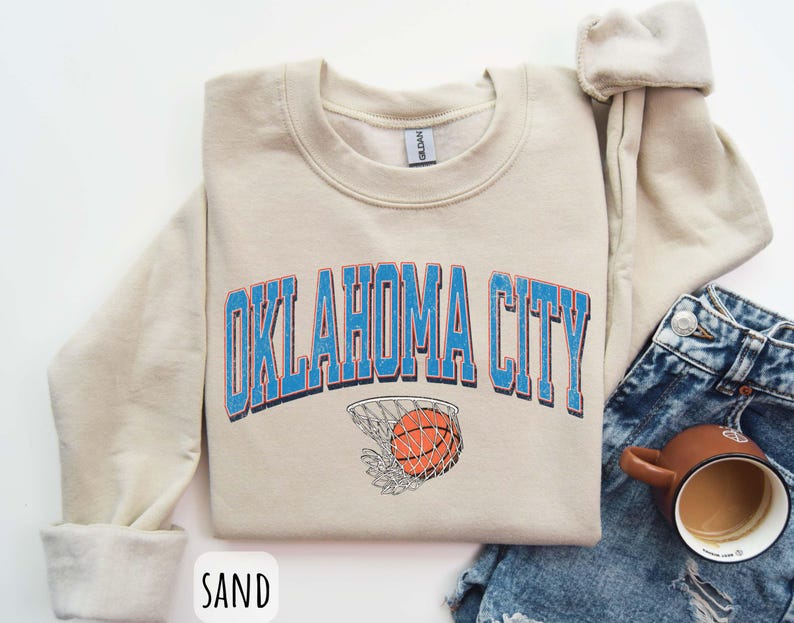 May include: A cream coloured sweatshirt with the text "OKLAHOMA CITY" in blue letters with a basketball and net graphic. The sweatshirt is on a denim background.
