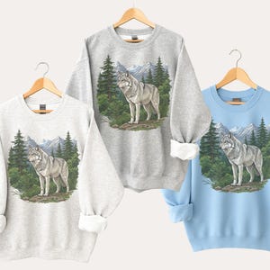 Op de afbeelding: Drie sweatshirts in lichtgrijs, grijs en lichtblauw, elk met een wolfillustratie. De wolf wordt afgebeeld in een boslandschap met bergen op de achtergrond. De sweatshirts worden op houten hangers getoond.