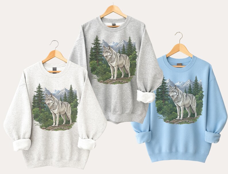Op de afbeelding: Drie sweatshirts in lichtgrijs, grijs en lichtblauw, elk met een wolfillustratie. De wolf wordt afgebeeld in een boslandschap met bergen op de achtergrond. De sweatshirts worden op houten hangers getoond.