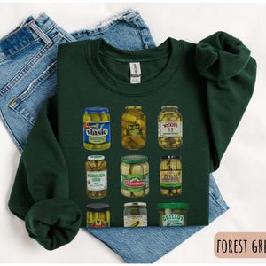 Op de afbeelding: Bostgroen sweatshirt met een afbeelding van verschillende augurkenpotten. De potten zijn gevuld met augurken en hebben verschillende merknamen. Het sweatshirt ligt op een blauwe spijkerbroek.