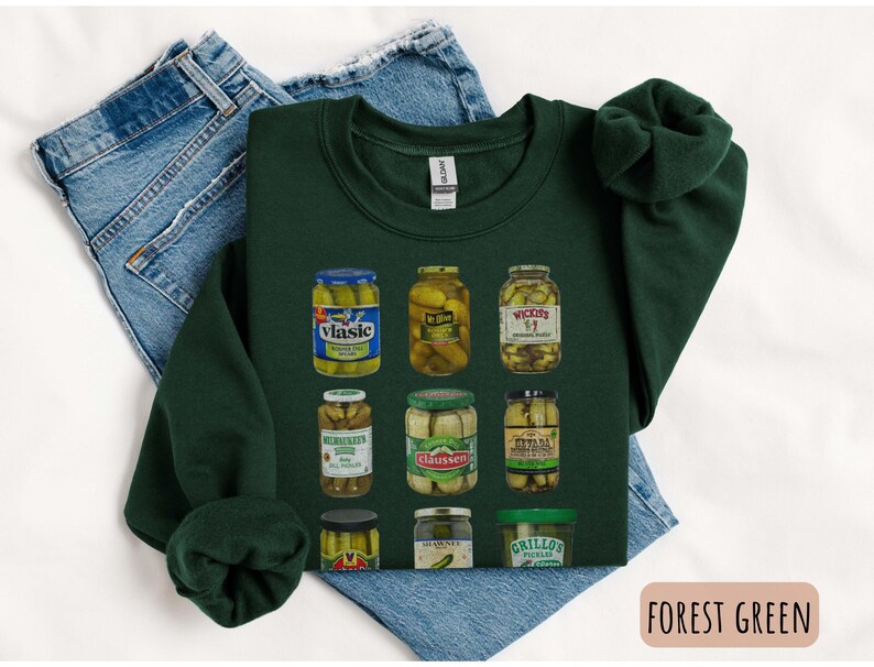 Op de afbeelding: Bostgroen sweatshirt met een afbeelding van verschillende augurkenpotten. De potten zijn gevuld met augurken en hebben verschillende merknamen. Het sweatshirt ligt op een blauwe spijkerbroek.