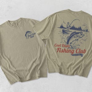 May include: Two beige t-shirts. One has a small blue graphic of a fish and the text "Reel Cool Dad." The other shirt has a larger graphic of a fisherman, a fish jumping out of the water, and the text "Cool Dads Fishing Club."