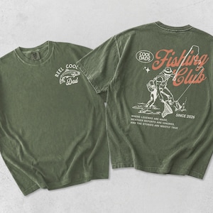 May include: Two olive green t-shirts. One has a white fish graphic and the text "REEL COOL Dad". The other features a fishing scene graphic with the text "COOL DADS Fishing Club SINCE 2026".
