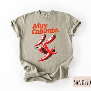 May include: Sandstone-colored t-shirt with the words "Muy Caliente" in red, above a graphic of three red chili peppers. The shirt is a relaxed fit, with rolled-up sleeves.