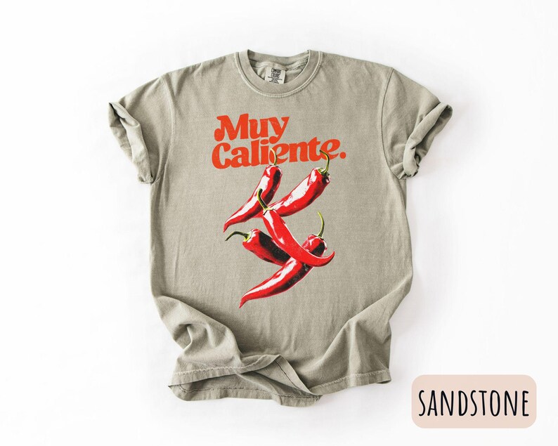 May include: Sandstone-colored t-shirt with the words "Muy Caliente" in red, above a graphic of three red chili peppers. The shirt is a relaxed fit, with rolled-up sleeves.