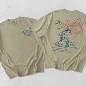 May include: Two beige t-shirts with fishing-themed designs. One displays a blue fish graphic with "REEL COOL Dad" text. The other has a fisherman graphic with "Fishing Club" and "SINCE 2026" text.