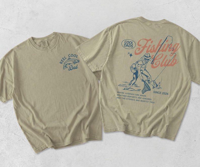 May include: Two beige t-shirts with fishing-themed designs. One displays a blue fish graphic with "REEL COOL Dad" text. The other has a fisherman graphic with "Fishing Club" and "SINCE 2026" text.
