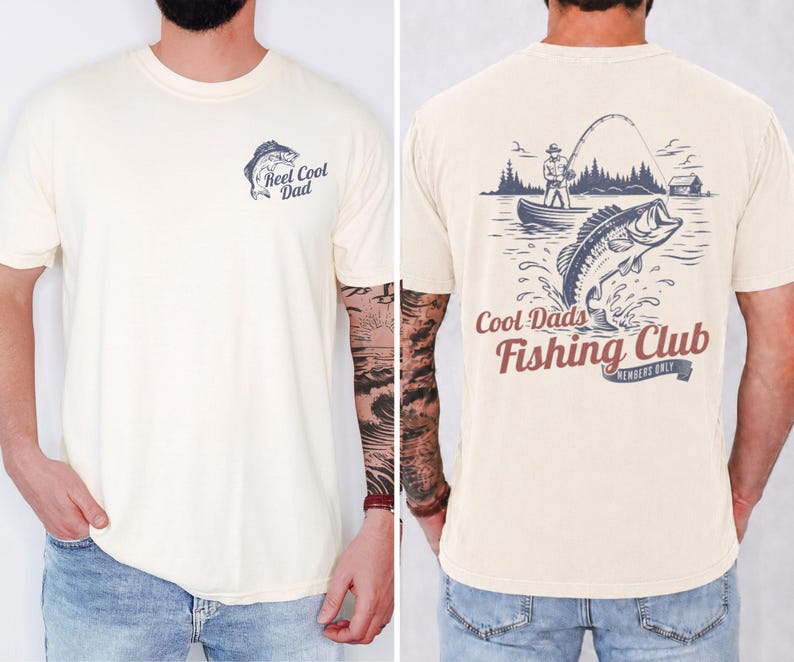 May include: Cream-colored t-shirt with a fishing-themed graphic. The front features a fish and the text "Reel Cool Dad." The back displays a fisherman in a boat, a jumping fish, and the words "Cool Dads Fishing Club."
