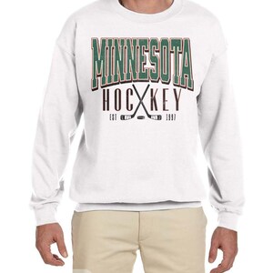 Peut inclure: Sweat-shirt blanc avec un imprim&eacute; graphique vert et rouge qui dit "Minnesota Hockey Est 1997" avec des b&acirc;tons de hockey crois&eacute;s et une rondelle.