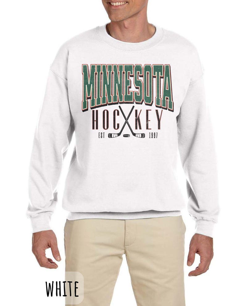 Peut inclure: Sweat-shirt blanc avec un imprim&eacute; graphique vert et rouge qui dit "Minnesota Hockey Est 1997" avec des b&acirc;tons de hockey crois&eacute;s et une rondelle.