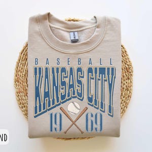 Peut inclure: Un sweat-shirt beige avec le texte "BASEBALL KANSAS CITY 19 69" en lettres bleues et dor&eacute;es. Le texte est entour&eacute; d'un ballon de baseball et de chauves-souris crois&eacute;es.